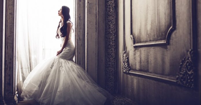 Bride in a white wedding dress standing beside a bright window in an ornate room.