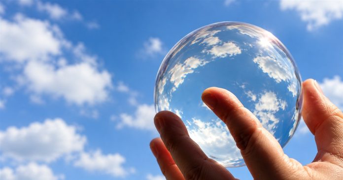 A hand holds a clear crystal ball against blue sky and white clouds.